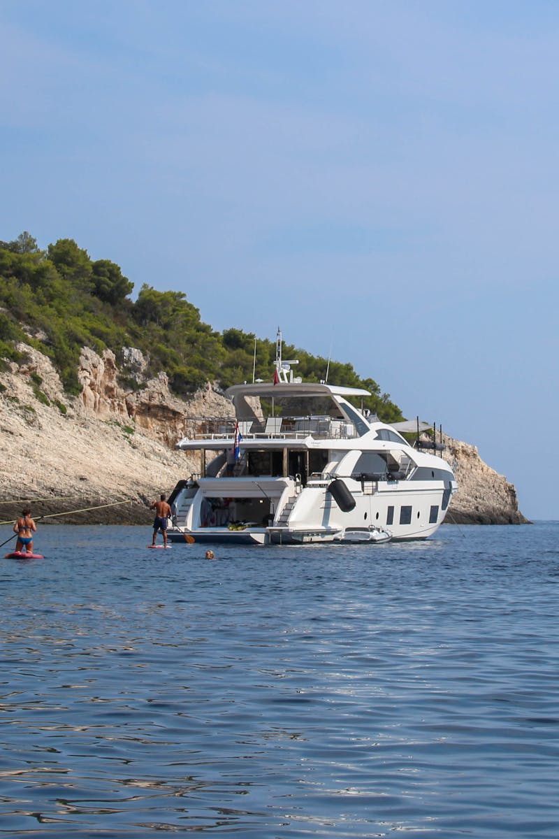 Luxury yachts anchored by a scenic rocky shoreline with people swimming and enjoying the sea.